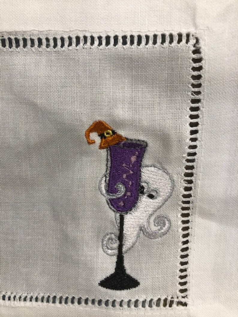 May include: White linen cocktail napkin with an embroidered Halloween design. The design features a purple cocktail glass with a witch's hat, and a friendly ghost. The napkin has a decorative stitched border.