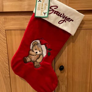 May include: Red Christmas stocking with a white cuff embroidered with the name "Sawyer" in burgundy script. A tag reads "Love Mom & Dad 2025". The stocking features a teddy bear wearing a Santa hat.