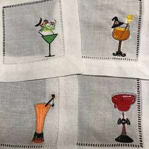May include: Set of four white linen cocktail napkins with embroidered Halloween-themed drink designs. Each napkin features a different cocktail glass with a unique design, including a witch's hat, broom, and bat.
