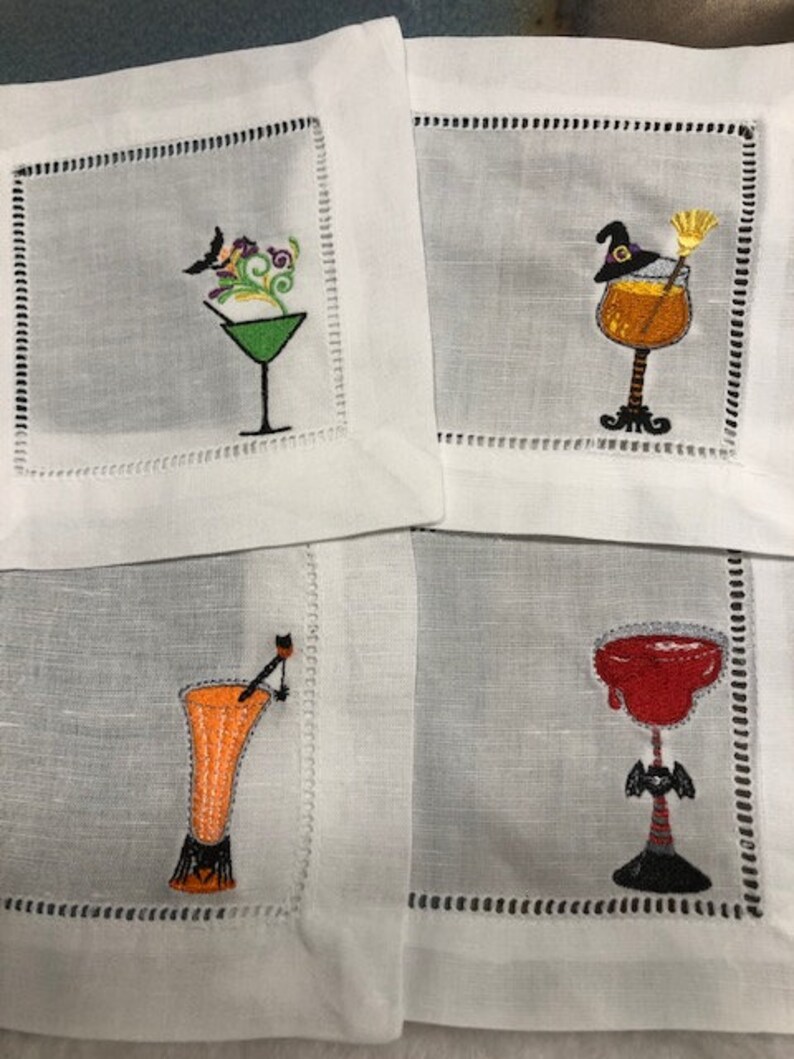 May include: Set of four white linen cocktail napkins with embroidered Halloween-themed drink designs. Each napkin features a different cocktail glass with a unique design, including a witch's hat, broom, and bat.