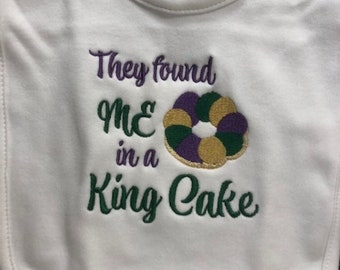 May include: White baby bib with green, purple, and gold embroidery that reads "They found me in a King Cake".