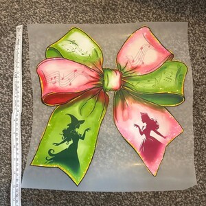 May include: A large, colourful bow design with green, pink, and red hues. The bow features musical notes and silhouettes of a witch and a princess. The bow is on a translucent background.