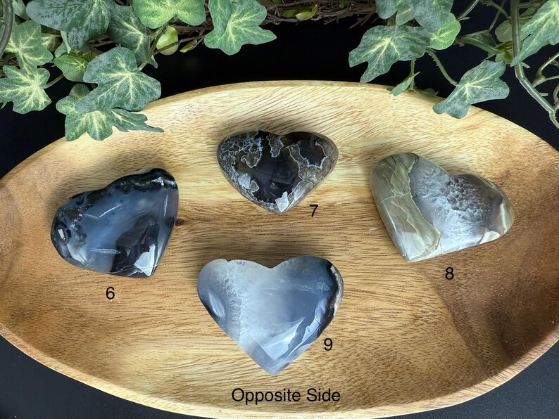 May include: Four heart-shaped stones in shades of gray, white, and black are displayed on a wooden tray. The stones have unique patterns and textures. The tray is surrounded by green foliage.