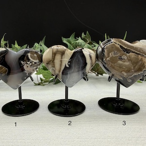 May include: Three heart-shaped decorative objects made of polished stone, each mounted on a black metal stand. The stones feature unique patterns in shades of gray, brown, and cream. The stands have a circular base.