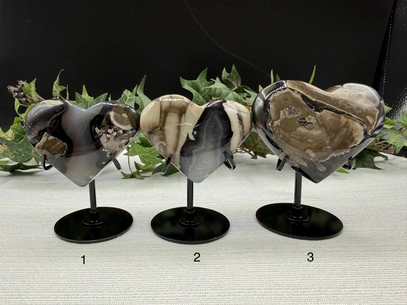 May include: Three heart-shaped decorative objects made of polished stone, each mounted on a black metal stand. The stones feature unique patterns in shades of gray, brown, and cream. The stands have a circular base.