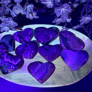 May include: A collection of heart-shaped stones in shades of purple and violet, arranged on a wooden plate. The stones have a marbled appearance and are illuminated with a purple light, creating a glowing effect. Decorative ivy is in the background.