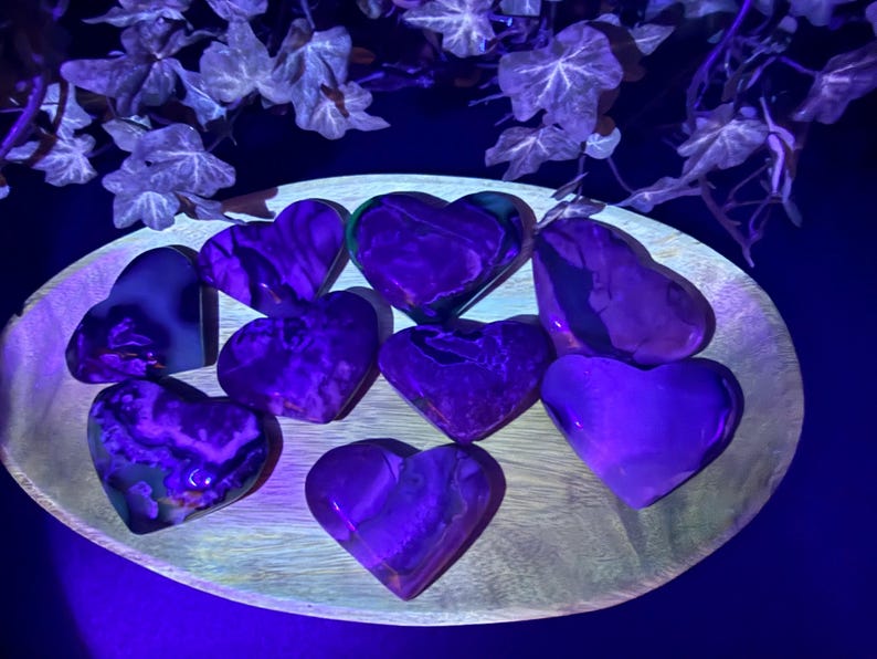 May include: A collection of heart-shaped stones in shades of purple and violet, arranged on a wooden plate. The stones have a marbled appearance and are illuminated with a purple light, creating a glowing effect. Decorative ivy is in the background.