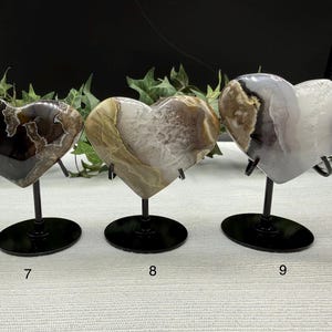 May include: Three heart-shaped agate stones on black metal stands. The stones vary in color, with patterns of brown, white, and gray. Each heart is approximately 4 inches wide. The stands have a circular base and a curved support.