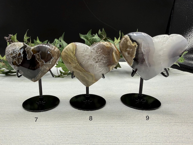 May include: Three heart-shaped agate stones on black metal stands. The stones vary in color, with patterns of brown, white, and gray. Each heart is approximately 4 inches wide. The stands have a circular base and a curved support.