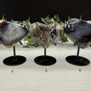 May include: Three heart-shaped stone sculptures on black metal stands. The stones have unique patterns in shades of gray, brown, and white. The stands are on a light-colored surface, with greenery in the background. Numbers 4, 5, and 6 are visible.