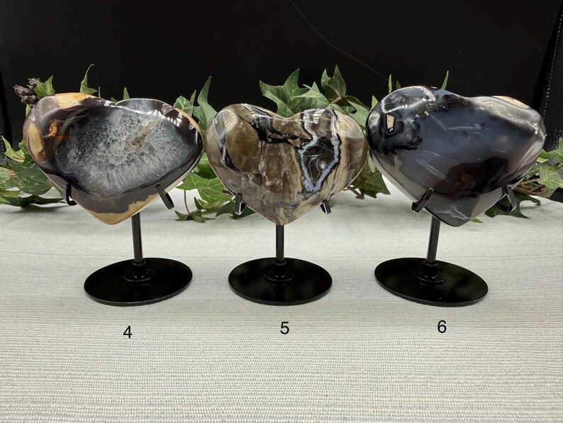 May include: Three heart-shaped stone sculptures on black metal stands. The stones have unique patterns in shades of gray, brown, and white. The stands are on a light-colored surface, with greenery in the background. Numbers 4, 5, and 6 are visible.