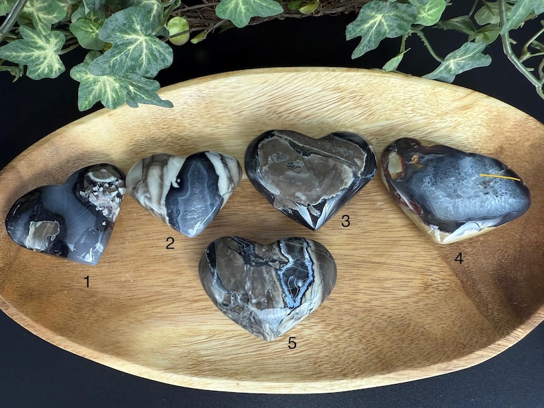 May include: Five heart-shaped stones with unique patterns and colors, displayed in a wooden tray. The stones vary in shades of gray, black, and brown, with visible veining and textures. The tray is oval-shaped and made of light-colored wood.
