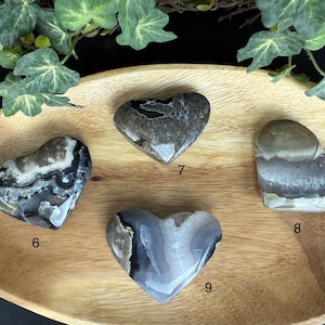 May include: Four heart-shaped stones with unique patterns and colors. The stones are shades of gray, white, and brown. They are displayed on a wooden tray with green foliage in the background. Each stone is numbered.