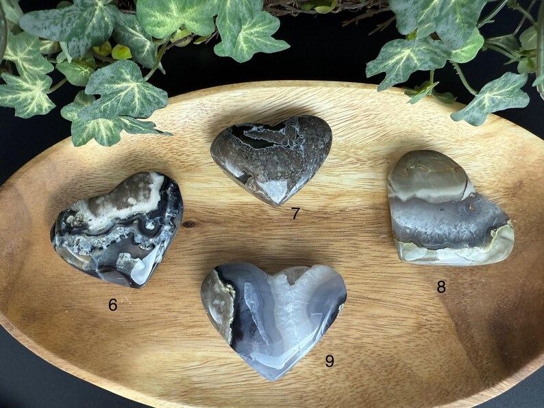 May include: Four heart-shaped stones with unique patterns and colors. The stones are shades of gray, white, and brown. They are displayed on a wooden tray with green foliage in the background. Each stone is numbered.