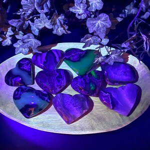 May include: A collection of heart-shaped stones in shades of purple, green, and white, arranged on a wooden, oval-shaped tray. The stones have a marbled appearance and are illuminated with a blue light, creating a glowing effect. Decorative ivy is in the background.