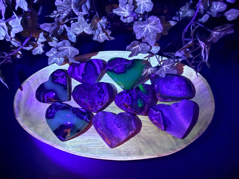 May include: A collection of heart-shaped stones in shades of purple, green, and white, arranged on a wooden, oval-shaped tray. The stones have a marbled appearance and are illuminated with a blue light, creating a glowing effect. Decorative ivy is in the background.