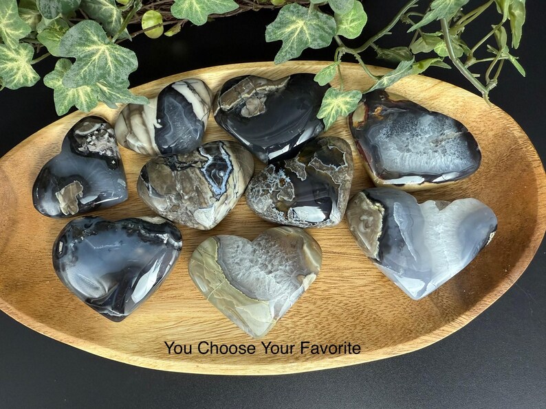 May include: A collection of heart-shaped stones in shades of gray, black, and white, displayed on a wooden tray. The stones have unique patterns and textures. The text "You Choose Your Favorite" is at the bottom.