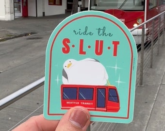 May include: A teal blue sticker with a white cartoon bird sitting on top of a red Seattle Transit streetcar. The sticker has the text "ride the S-L-U-T" in white letters.