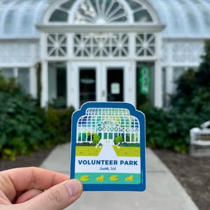 May include: A blue and green sticker with a white illustration of a greenhouse and the text "VOLUNTEER PARK Seattle, WA". There are four yellow ducklings swimming in a pond in front of the greenhouse.