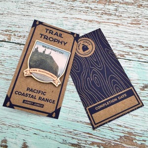 May include: A wooden trail trophy pin with a picture of Cape Disappointment, Washington. The pin is brown with a blue and white image of the lighthouse and the text "TRAIL TROPHY", "WASHINGTON", "CAPE DISAPPOINTMENT", "PACIFIC COASTAL RANGE", and "JUNKY JUNCO".