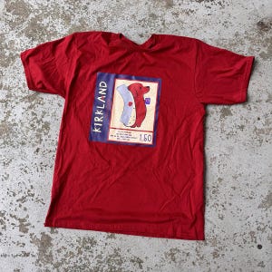 Puede incluir: Camiseta roja con un gráfico de un perrito caliente salchicha rojo en un bollo blanco, sobre un rectángulo azul con la palabra "KIRKLAND". El gráfico también incluye el número "1,50".