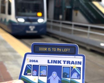 Puede incluir: Una pegatina azul y blanca con el texto "DOOR IS TO MY LEFT" y "LINK TRAIN". La pegatina presenta un tren de dibujos animados con un pingüino y otras aves en las ventanas. El número del tren es 206A.