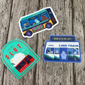 May include: Three colorful stickers featuring illustrations of public transportation. The first sticker is a teal blue bus with the text "County Transit" and "Seattle" on the front. The second sticker is a teal blue train with the text "ride the S-L-U-T" and a penguin on top. The third sticker is a blue train with the text "DOOR IS TO MY LEFT" and "LINK TRAIN" on the front.