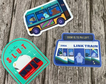 May include: Three colorful stickers featuring illustrations of public transportation. The first sticker is a teal blue bus with the text "County Transit" and "Seattle" on the front. The second sticker is a teal blue train with the text "ride the S-L-U-T" and a penguin on top. The third sticker is a blue train with the text "DOOR IS TO MY LEFT" and "LINK TRAIN" on the front.