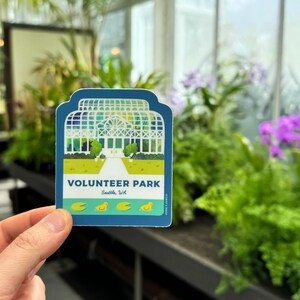 May include: A blue and green sticker with a white illustration of a greenhouse and the text "VOLUNTEER PARK Seattle, WA". There are four yellow ducklings and three green lily pads on a blue pond below the greenhouse.