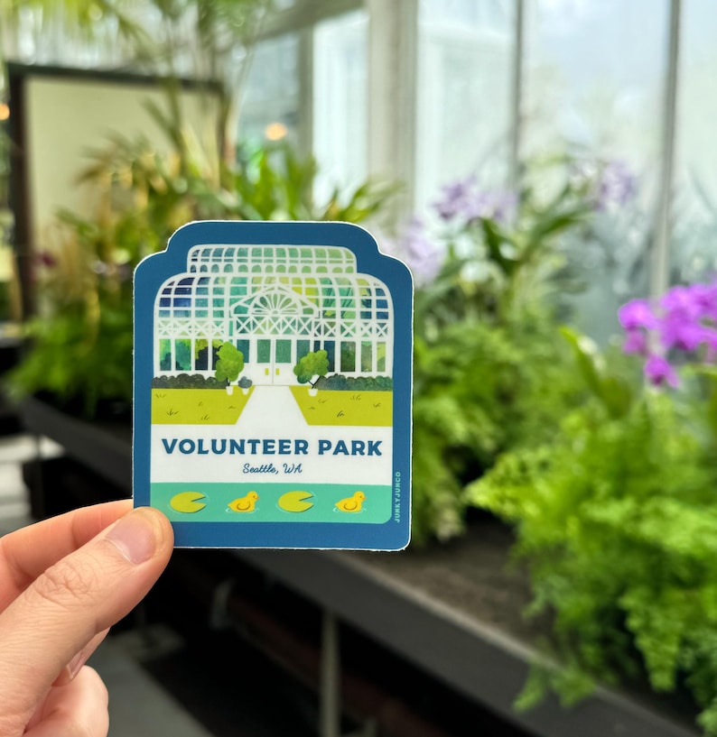 May include: A blue and green sticker with a white illustration of a greenhouse and the text "VOLUNTEER PARK Seattle, WA". There are four yellow ducklings and three green lily pads on a blue pond below the greenhouse.