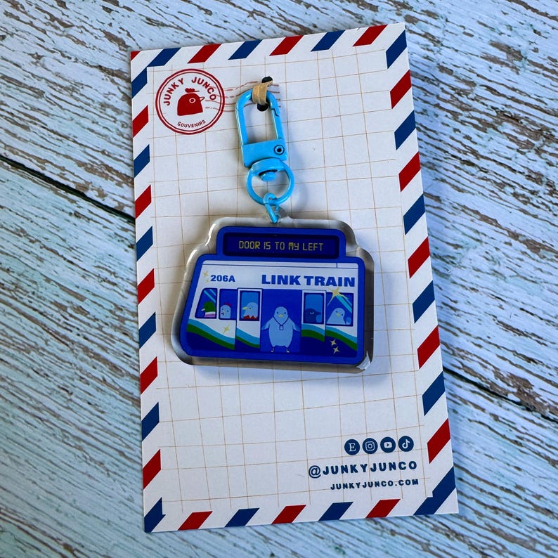 Puede incluir: Llavero de acr&iacute;lico azul y blanco con un ping&uuml;ino de dibujos animados montando un Link Train con el texto "DOOR IS TO MY LEFT" y "LINK TRAIN". El llavero tambi&eacute;n incluye un cierre de metal azul.