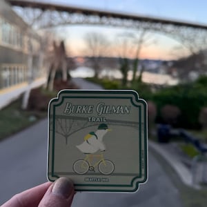 Puede incluir: Una pegatina con borde verde y las palabras "Burke Gilman Trail" y "Seattle, WA". La pegatina presenta un pato de dibujos animados montando en bicicleta amarilla. El fondo muestra un puente y una masa de agua.
