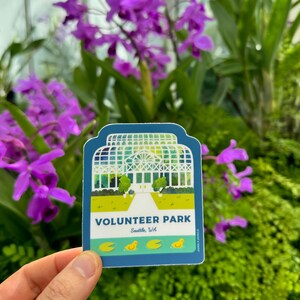 May include: A blue and green sticker with an illustration of a white greenhouse with a walkway leading to the entrance. The text "VOLUNTEER PARK" is printed in white on a green background. Below the text is "Seattle, WA" in white on a blue background. There are four yellow duck illustrations at the bottom of the sticker.