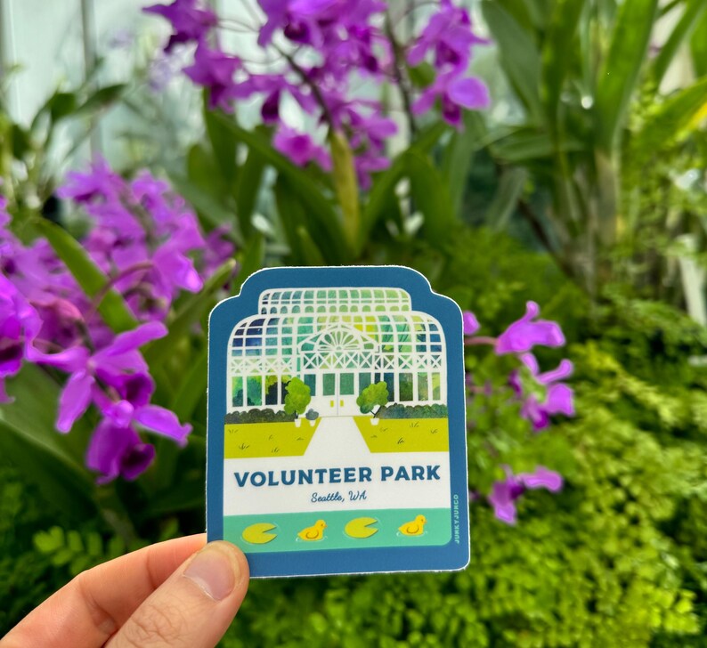 May include: A blue and green sticker with an illustration of a white greenhouse with a walkway leading to the entrance. The text "VOLUNTEER PARK" is printed in white on a green background. Below the text is "Seattle, WA" in white on a blue background. There are four yellow duck illustrations at the bottom of the sticker.