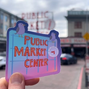 Puede incluir: Una pegatina rosa y azul con un patrón de cuadrícula blanca y dos pájaros blancos. La pegatina dice "Public Market Center" en letras amarillas y rojas.