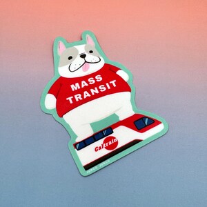 Puede incluir: Una pegatina troquelada con un bulldog de dibujos animados que lleva una camiseta roja con la inscripción "MASS TRANSIT". El bulldog está de pie sobre un tren Caltrain. La pegatina tiene un borde verde claro y el texto "JUNCO JUNCO" en la parte inferior.