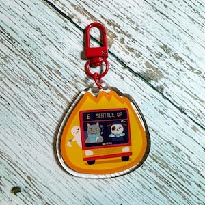 May include: A yellow and red acrylic keychain featuring a cartoon bus with a cat, a panda, and a bird on it. The bus has the text "E Seattle, WA" on the side. The keychain has a red metal clasp.