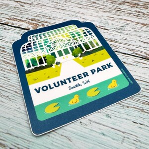 May include: A blue and green sticker with a watercolor illustration of a greenhouse and the text "Volunteer Park, Seattle, WA". There are three yellow rubber duckies and three green lily pads on a pond in front of the greenhouse.
