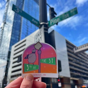以下が含まれることがあります： 「3rd Ave」と「Pine St」の標識に止まっている漫画のハトが描かれたステッカー。場所を示す「Seattle, WA」のタグ付き。ステッカーは、夕焼けをテーマにしたカラフルな背景が特徴です。