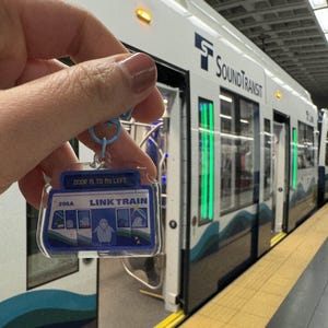 Puede incluir: Un llavero azul y blanco de Link Train con el texto "DOOR IS TO MY LEFT" y "LINK TRAIN" en &eacute;l. El llavero tambi&eacute;n tiene el n&uacute;mero "206A" y un gr&aacute;fico de ping&uuml;ino. El llavero se est&aacute; sosteniendo frente a un tren ligero de Sound Transit.