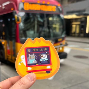 May include: A yellow and orange sticker with a cartoon bus on it. The bus is red and has a white sign that says "E Seattle, WA". There are three cartoon animals on the bus: a white bird, a gray cat, and a brown dog. The sticker is in front of a red bus.