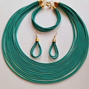 May include: A teal jewelry set featuring a multi-strand necklace, bracelet, and teardrop earrings. The necklace and bracelet have gold-colored clasps. The earrings have gold-colored caps. The set is displayed on a white surface.