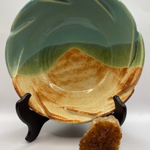May include: A large, ceramic bowl with a light blue glaze on the top half and a brown and yellow glaze on the bottom half. The bowl has a wavy rim and is sitting on a wooden stand.