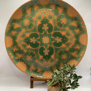 May include: A large, round ceramic plate with a green and orange geometric pattern. The plate is on a wooden stand and a small green plant is in front of it.