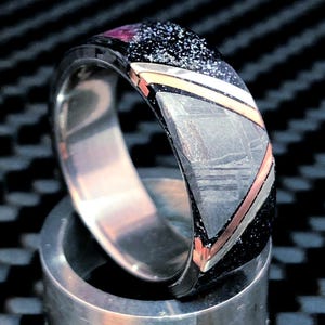 May include: A ring featuring a polished metallic inner band and an outer inlay composed of a dark, speckled material, a triangular grey textured segment, and thin diagonal bands of rose gold and silver metal, creating a multi-layered appearance.