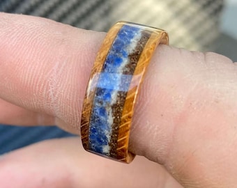 May include: A wooden ring with a band of blue and brown stone inlay. The ring is made of light brown wood and features a polished finish. The stone inlay is a mix of blue and brown hues.