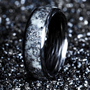 May include: A close-up of a black ring with a white and gray speckled inlay. The ring has a polished, reflective surface and sits on a dark, textured background. The ring is a piece of jewelry.