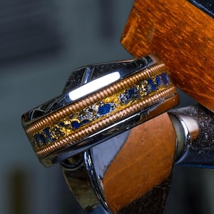 Puede incluir: Primer plano de un anillo plateado con una banda decorativa. La banda presenta un diseño de alambre de cobre, detalles dorados e incrustaciones de piedras azules. El anillo se encuentra sobre un fondo borroso.