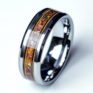 Puede incluir: Un anillo plateado con una banda central dorada e incrustaciones de piedras preciosas de colores. Las piedras preciosas son una mezcla de tonos naranja, rosa y morado. El anillo tiene una superficie pulida y reflectante, y las piedras están engastadas en una resina transparente.