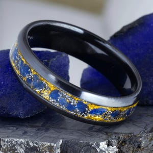 Könnte beinhalten: Ein schwarzer Bandring mit einer zentralen Einlage aus zerkleinertem blauem Stein, metallischen Silberflocken und goldfarbenen Blattakzenten. Der Ring hat eine glatte, polierte Oberfläche und ein abgerundetes Profil mit einem starken Kontrast.